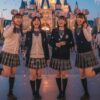 東京ディズニーリゾートのシンデレラ城前で制服を着てポーズを取る4人の日本人女性たち。夕暮れ時で、花火が上がっており、笑顔で楽しんでいる様子が伝わる。