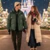 雪が舞う冬の夜のディズニーランドで、暖かそうなダウンジャケットを着て手をつなぎ、笑顔で見つめ合う日本人カップル。背景にはライトアップされたシンデレラ城とクリスマスツリーが輝いている。