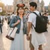 ディズニーランドのシンデレラ城を背景に、笑顔で手をつなぐ日本人カップル。男性は黒いリュックサックを背負い、女性はトートバッグとミニーの耳カチューシャをつけている。