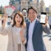 東京ディズニーランドのシンデレラ城を背景に、笑顔で株主優待パスポートと株価チャートが表示されたスマートフォンを持つ日本人夫婦。二人の頭上には、株主優待の廃止や変更に関する疑問や不安を表す吹き出しが描かれている。